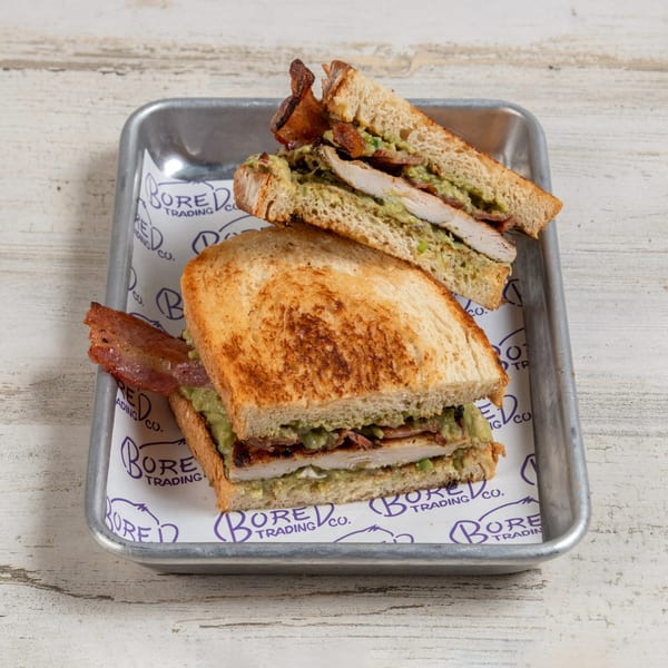 Grilled chicken and avocado sandwich with bacon on toasted bread, served on a metal tray.